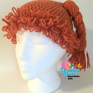 May include: A crocheted orange wig hat with a braided ponytail. The hat has a textured design and fringe around the brim. The ponytail is secured with a white ribbon. The hat is displayed on a white mannequin head.