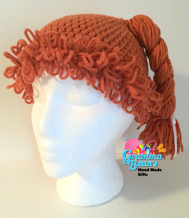 May include: A crocheted orange wig hat with a braided ponytail. The hat has a textured design and fringe around the brim. The ponytail is secured with a white ribbon. The hat is displayed on a white mannequin head.