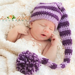 May include: A newborn baby wearing a purple and lavender striped crochet hat with a long, pointed tail and a large pom-pom. The baby is wrapped in a soft, white blanket. The image is from Amanda Hobbs Photography.