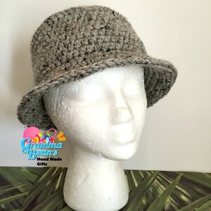 May include: A grey crocheted bucket hat with a wide brim. The hat is on a white mannequin head. The background is a green and brown tropical leaf pattern.