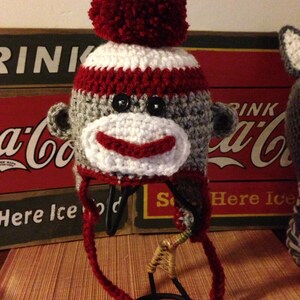 May include: A crocheted monkey hat with a red pom-pom on top. The hat is gray, white, and red and has a monkey face with black eyes.