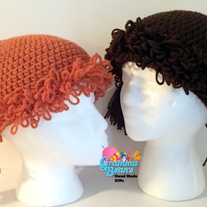 May include: Two crochet hats resembling pigtails, one in orange and the other in brown, are displayed on white mannequin heads. The orange hat features a braided pigtail and a white ribbon. The brown hat has a ponytail. The hats are handmade.