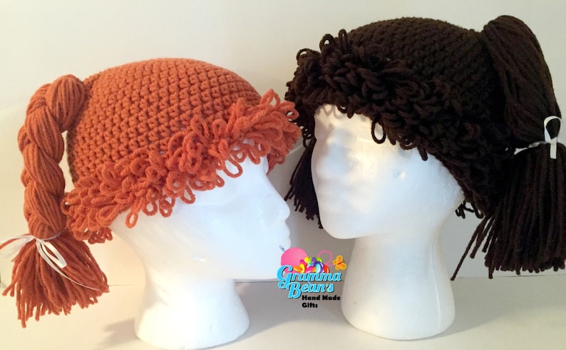 May include: Two crochet hats resembling pigtails, one in orange and the other in brown, are displayed on white mannequin heads. The orange hat features a braided pigtail and a white ribbon. The brown hat has a ponytail. The hats are handmade.