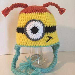 May include: A yellow, orange, and blue crocheted hat with a single eye and a black strap. The hat has two long braids on top and a long braided strap with a blue pom-pom at the end.