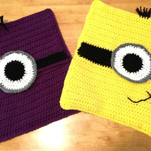 May include: Two crocheted Minion-themed pillows, one purple and one yellow, with black eye patches and a single eye on each. The pillows are on a wooden surface.