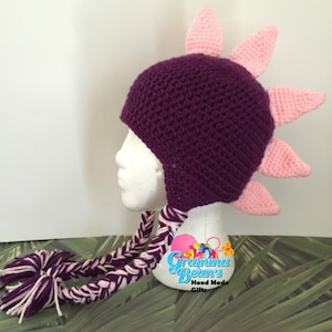 May include: Purple and pink crocheted hat with a spiky design and long braided ties. The hat is on a white mannequin head. The hat is labeled "Gramma Bean's Hand Made Gifts".