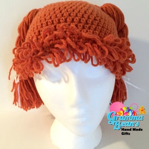 May include: A handmade orange crochet hat with fringe and braided yarn details. The hat is displayed on a white mannequin head. The hat has the text "Gramma Bean's Hand Made Gifts" in the lower right corner.