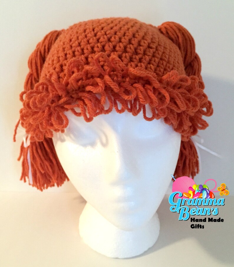 May include: A handmade orange crochet hat with fringe and braided yarn details. The hat is displayed on a white mannequin head. The hat has the text "Gramma Bean's Hand Made Gifts" in the lower right corner.