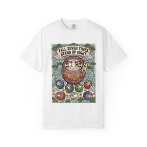 Könnte beinhalten: Weißes T-Shirt mit einem farbenfrohen Aufdruck einer Daruma-Puppe und kleineren Daruma-Puppen. Der Aufdruck enthält den Text "FALL SEVEN TIMES, STAND UP EIGHT" in einem grünen Banner. Die Daruma-Puppe ist rot mit schwarzen Augen und dem Wort "福" auf dem Bauch.