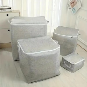 May include: A set of four square storage bins in varying sizes, crafted from a light gray fabric with a subtle textured pattern. Each bin features a white zippered lid and two side handles, providing a structured and uniform look for organization.