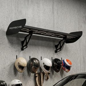May include: A black Porsche rear wing mounted on a gray concrete wall, above a wooden shelf. Below the shelf are several racing helmets in various colors, including white, black, and orange. A black sports car is parked below.