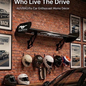 Puede incluir: Decoración para entusiastas de los coches con un alerón Mustang negro, carteles de coches antiguos enmarcados y cascos de carreras en una pared de ladrillo. El texto dice "Built For Those Who Live The Drive."