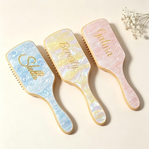 May include: Three rectangular hairbrushes with a pearlescent finish in blue, yellow, and pink. Each brush features a gold script name printed on the handle: Stella, Beverly, and Galina. The brushes have light-colored wooden handles and sides.