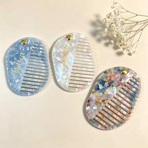 Peut inclure: Trois peignes à cheveux ovales en acrylique marbré. Un peigne est bleu clair, un est blanc irisé et un présente un motif confettis multicolore. Chaque peigne possède un rivet en métal doré et un nom gravé en écriture cursive dorée.