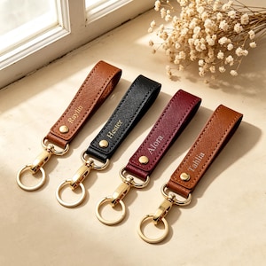 May include: Four leather keychains with gold-tone metal hardware and circular rings. Each strap features a textured finish and a gold-stamped name: Raylin on tan, Hester on black, Alora on burgundy, and Tahlia on brown leather. Each has a snap button.