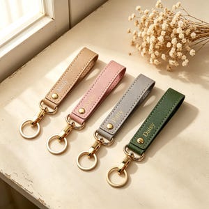 May include: Four leather keychains in beige, pink, gray, and dark green. Each strap features a gold-tone metal snap button, a lobster clasp, and a circular ring. The straps are personalized with the names Molly, Kate, Patty, and Daisy.
