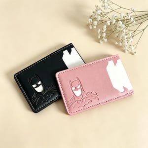 May include: Two rectangular card holders, one black and one pink, each featuring an embossed Batman logo on the front. Both items have white stitching along the edges and a white cutout section on the right side for card insertion and access.