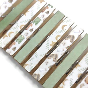 May include: A set of wooden clothespins with a green and white patterned design. The pattern features stars, moons, leaves, and small illustrations of baby items like booties, hats, and onesies.