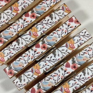 May include: A set of six wooden clothespins with a white background and a floral pattern featuring small birds. The birds are in shades of blue, yellow, and orange. The flowers are pink and red.