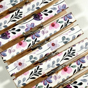 May include: A set of wooden clothespins with a white background and a floral pattern in shades of pink, purple, and gray. The clothespins are decorated with a watercolor-style floral design.