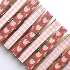 May include: A collection of wooden clothespins with decorative paper. The paper features a pattern of pink strawberries on a red background and pink gingham. The clothespins have silver metal springs.