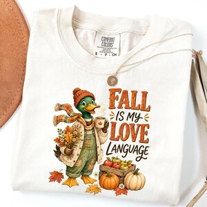 May include: A cream-colored t-shirt featuring a graphic of a duck wearing a knit hat, scarf, and overalls, holding a bouquet of autumn leaves and a mug. The text reads FALL IS MY LOVE LANGUAGE, accompanied by a wooden crate of apples and pumpkins.