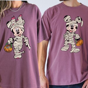 May include: Two mauve-colored short-sleeve t-shirts featuring graphic prints of cartoon characters wrapped in white bandages like mummies. One character wears a bow, and both hold orange jack-o'-lantern buckets filled with colorful candy.