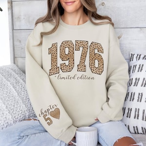 May include: A cream-colored crewneck sweatshirt featuring the large block numbers 1976 in a brown and black leopard print. Below the numbers, the text reads limited edition in a cursive font. The sleeve displays the text chapter 5 and a heart.