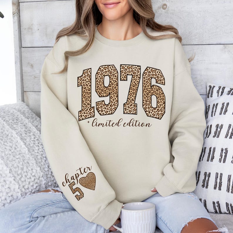 May include: A cream-colored crewneck sweatshirt featuring the large block numbers 1976 in a brown and black leopard print. Below the numbers, the text reads limited edition in a cursive font. The sleeve displays the text chapter 5 and a heart.