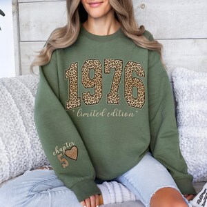 May include: An olive green crewneck sweatshirt featuring the year 1976 in a large leopard print font across the chest, with the text limited edition below it. The left sleeve displays the text chapter 5 with a leopard print heart icon.