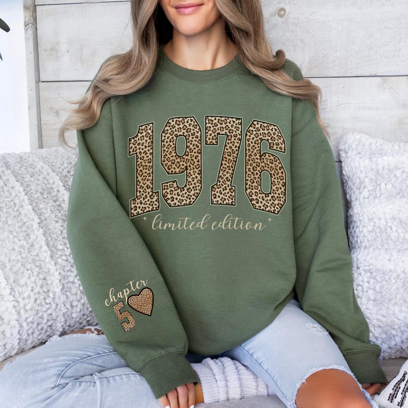 May include: An olive green crewneck sweatshirt featuring the year 1976 in a large leopard print font across the chest, with the text limited edition below it. The left sleeve displays the text chapter 5 with a leopard print heart icon.