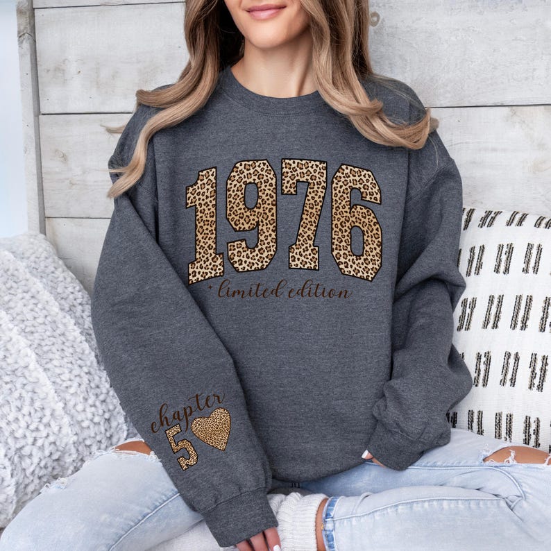 May include: A dark gray crewneck sweatshirt featuring the year 1976 in a large leopard print font with the text limited edition underneath. The left sleeve displays the text chapter 5 with a small leopard print heart icon below the number.