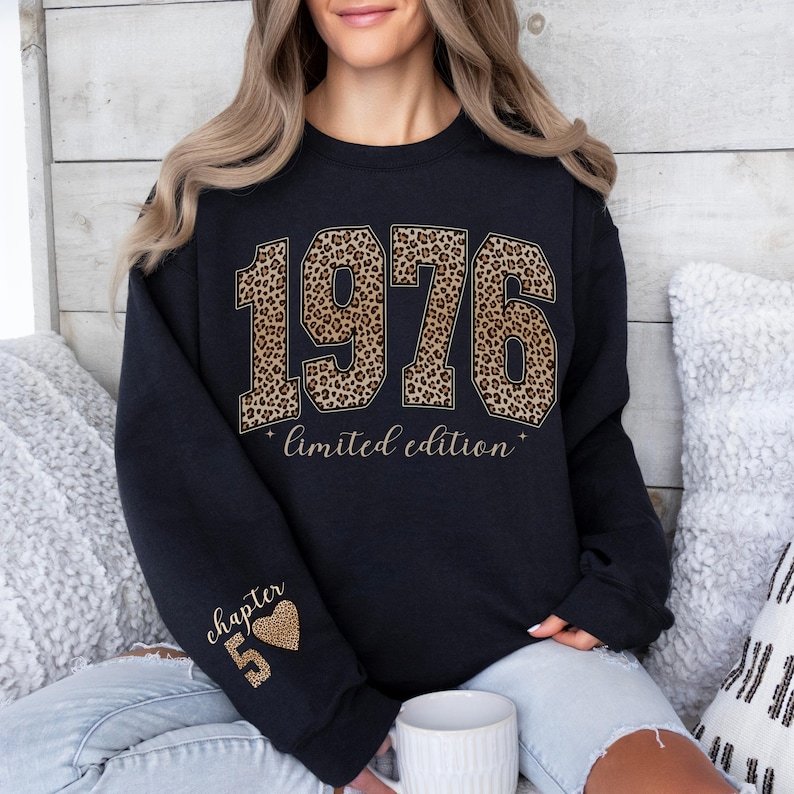 May include: A black crewneck sweatshirt featuring the text 1976 in a brown and black leopard print pattern, with the words limited edition printed below. The left sleeve displays the text chapter 5 with a small leopard print heart icon.