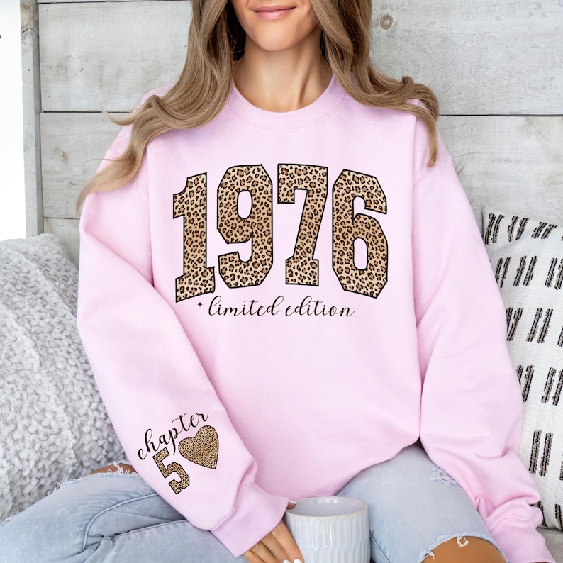 May include: A light pink crewneck sweatshirt featuring the year 1976 in a large leopard print font across the chest, with the text limited edition below it. The left sleeve displays the text chapter 50 next to a leopard print heart graphic.