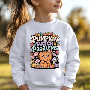 May include: A white long-sleeve sweatshirt featuring a colorful graphic print with the text PUMPKIN PATCH PROBLEMS. The design includes a central orange pumpkin character, various smaller pumpkins, mushrooms, autumn leaves, and candy pieces.