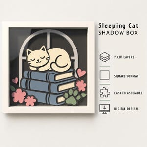 May include: A square white shadow box frame containing a layered paper art design of a cream-colored cat sleeping on a stack of three blue books in front of a window. The scene includes pink hearts, pink flowers, and a green paw print icon.