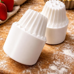 May include: Two white pie crust weights with a ridged design sit on a wooden cutting board dusted with flour. Fresh strawberries and a pie crust are in the background. Baking tools for making pies.
