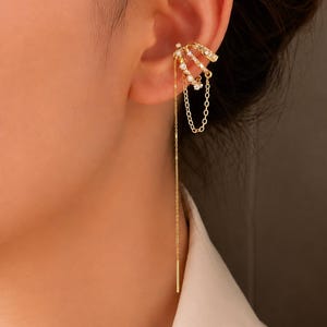 May include: A gold-toned ear cuff featuring three stacked bands adorned with small clear stones. Two of the bands are connected by a delicate gold chain, and a long, thin gold chain drapes downward from the cuff, ending in a straight metal bar.