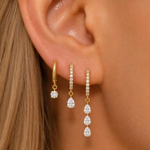 May include: Three gold hoop earrings worn in a row. The first is a plain gold hoop with a single round clear stone charm. The second is a pavé hoop with a pear-shaped stone charm. The third is a pavé hoop with three dangling pear-shaped stones.
