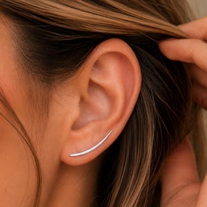 May include: A sleek, curved silver ear climber earring that follows the natural contour of the earlobe. The earring features a smooth, polished metallic finish and a minimalist, elongated arc shape that extends upward along the lower ear cartilage.