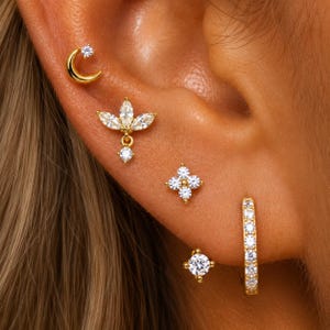 May include: A set of five gold-tone earrings featuring a crescent moon stud with a clear stone, a marquise-cut floral cluster stud with a dangling stone, a four-stone flower stud, a single round stone stud, and a small hoop earring lined with clear stones.