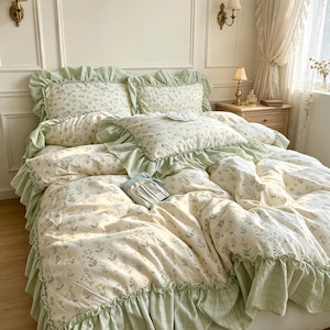 Könnte beinhalten: Ein Bettwäsche-Set bestehend aus einem cremefarbenen Bettbezug und Kissenbezügen mit einem zarten Blumenmuster in Grün- und Gelbtönen. Die Ränder sind mit einem breiten, gerüschten hellgrünen Stoffbesatz versehen, der für eine geschichtete Optik sorgt.