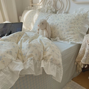 Könnte beinhalten: Ein Bettwäscheset bestehend aus einem cremefarbenen Bettbezug und Kissenbezügen mit zartem blauem Blumenmuster und weißen Rüschenkanten. Ein cremefarbenes Plüschkaninchen sitzt auf dem Bett, das mit einem hellblau-weiß karierten Laken bezogen ist.