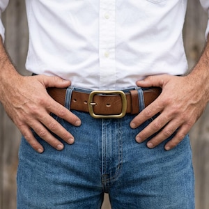 Peut inclure: Une ceinture en cuir marron avec une boucle rectangulaire en laiton portée dans les passants d'un jean bleu. La ceinture présente une finition lisse et une fermeture à ardillon unique, fixée autour de la taille sur une chemise blanche.