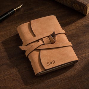 May include: A tan suede journal featuring a wrap-around strap closure with a knot detail. The front cover is embossed with the letters S and R separated by a small heart symbol. The journal has a rectangular shape and a soft, matte texture.