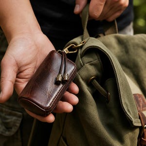 May include: A dark brown leather key case with a zipper closure, featuring two leather pull tabs with decorative metallic beads. The case is attached to a brass-colored metal swivel clasp, which is hooked onto the strap of an olive green canvas backpack.