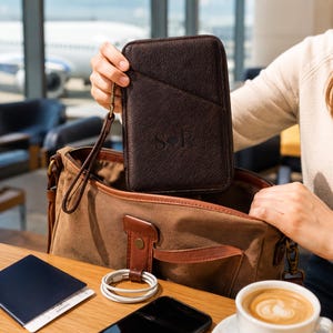 Könnte beinhalten: Eine dunkelbraune Reisebrieftasche aus strukturiertem Leder mit Handschlaufe und den geprägten Buchstaben S Herz P. Die Brieftasche wird über einer braunen Umhängetasche aus Segeltuch mit Lederakzenten, einem weißen Ladekabel und einem blauen Reisepass gehalten.