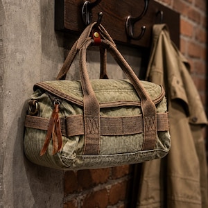 May include: A small olive green canvas duffel bag featuring brown woven fabric straps, a top handle with a brown leather accent, and a zipper closure with brown leather pull tabs. The bag has a structured rectangular shape with reinforced side panels.