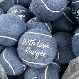 以下が含まれることがあります： 白い曲線状の縫い目と白い文字でWith love, Rangerとプリントされた、濃い青色のフェルト製テニスボールの集合体。ボールの毛羽立った質感がグループ全体に見られ、複数のボールが積み重なっている。
