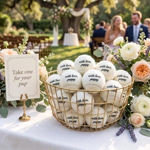 May include: A gold wire basket filled with white tennis balls, each printed with the black text "with love, poppy". Next to the basket is a white sign on a gold stand that reads "Take one for your pup" in black lettering, surrounded by floral arrangements.